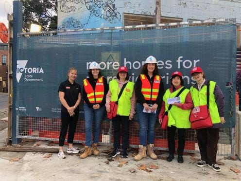 Seven women stand in front of construction siding. Three are from The Big Issue, wearing their caps, high-vis and satchel bags.