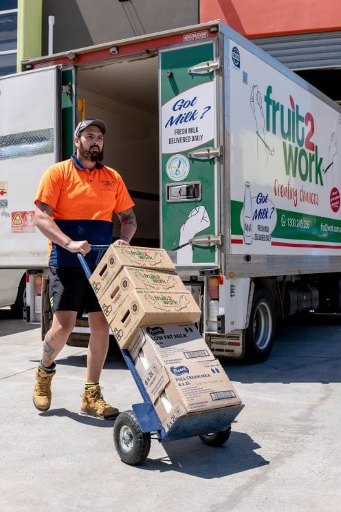 Man in orange high-viz t-shirt pushing kick-trolley loaded up with boxes, all in front of a truck bearing the logo of Fruit2Work.