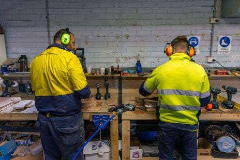 Two men facing away from camera with large earmuffs, both working at a woodworking table.