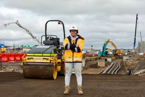 You woman at a construction site, standing proudly in front of double-drum roller with arms crossed.